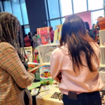 Deux visiteuses feuillettent des livres et explorent un stand coloré dédié à la littérature jeunesse et aux créations artisanales.