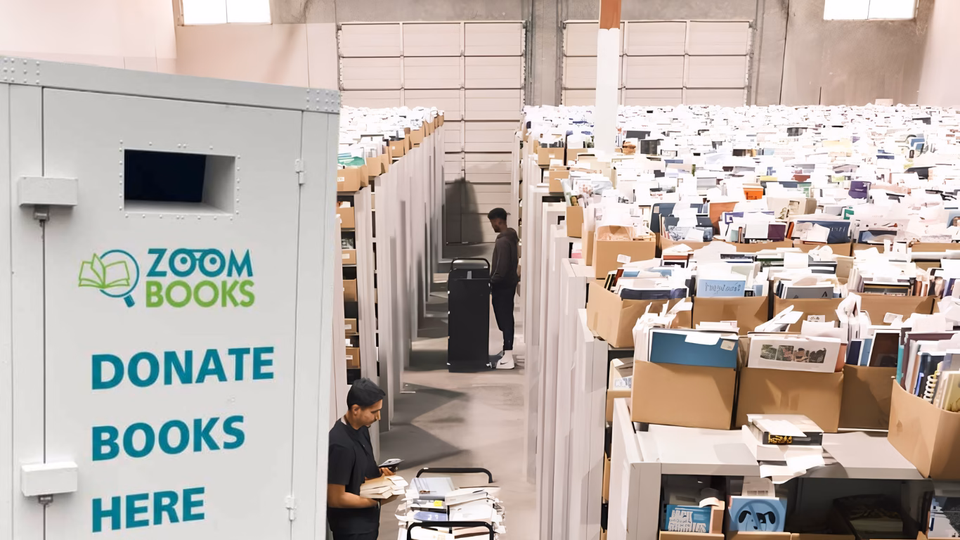Montage photo montrant au premier plan une borne de collecte de livres blanche estampillée du logo « Zoom Books » avec l'inscription « DONATE BOOKS HERE ». À l'arrière-plan, la borne s'ouvre sur un immense entrepôt industriel où s'alignent des rangées interminables de rayonnages surchargés de cartons et de livres d'occasion en vrac, au milieu desquels travaillent deux employés.